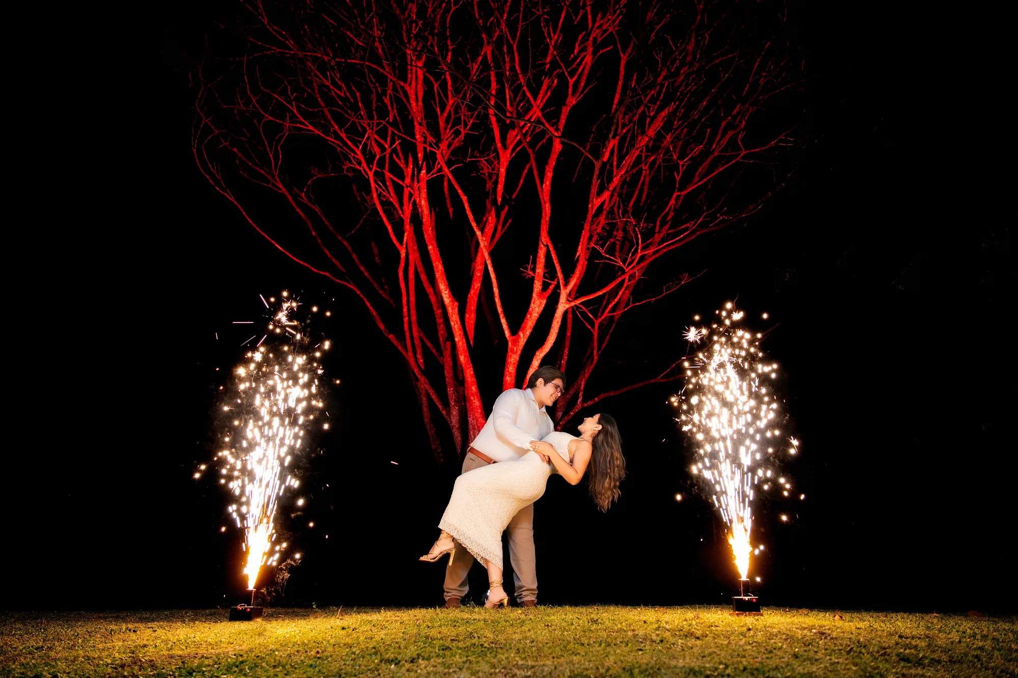 Los novios bailan entre dos fuentes de chispas bajo un árbol iluminado